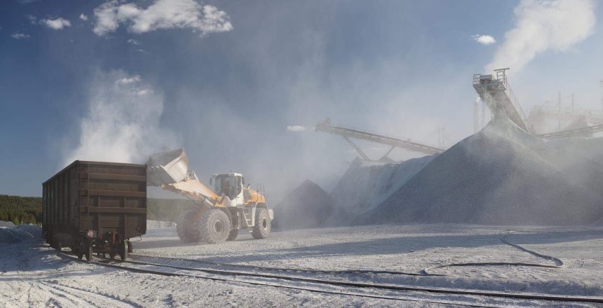 Front-end loader loads railway cars with crushed stone at an mining and processing plant in the background of stone crushing equipment in a cloud of stone dust, panorama.